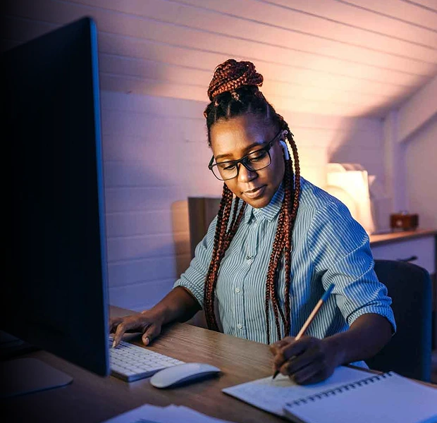 A woman in glasses writing into a notebook in front of her desktop